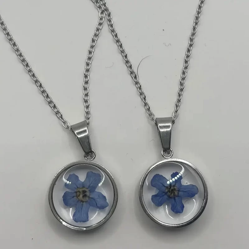 Silver-Plated Round Botanical Necklace with Blue Dried Flowers Two silver-plated round pendants with real blue dried flowers on matching silver chains, displayed on a white surface.