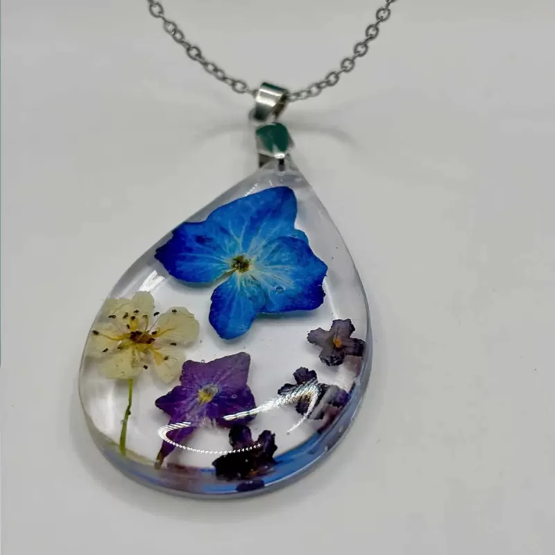 Druppelvormige blauwe geperste bloemen ketting met grote blauwe bloem, witte en paarse bloempjes in heldere hars aan zilverkleurige ketting