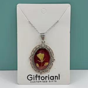 Handgemaakte droogbloemen ketting met geel bloemetje in rode epoxy en ovale roestvrijstalen hanger op merkkaart